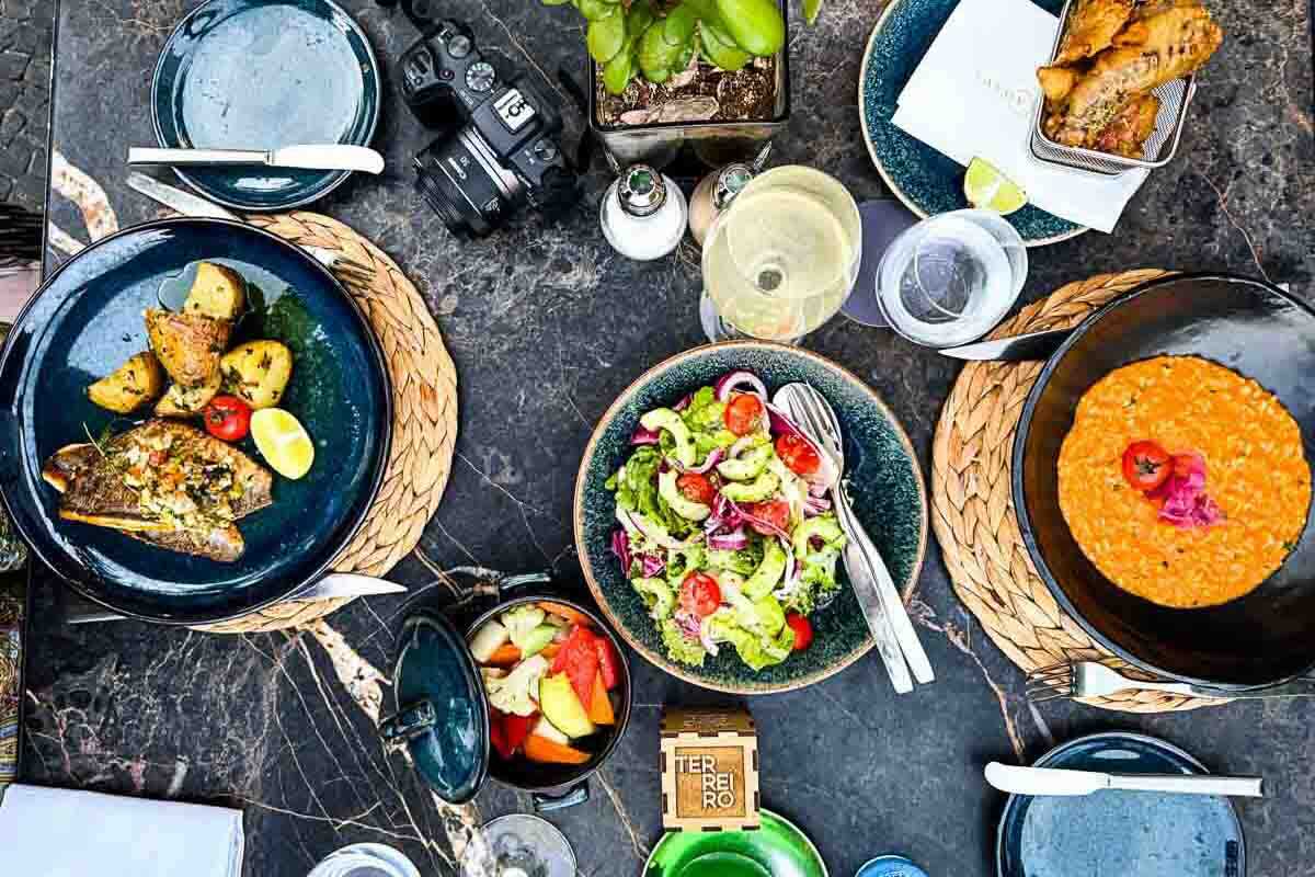 Overhead view of a table with plates of roasted potatoes, fish, salad, risotto, bread and a small plant.