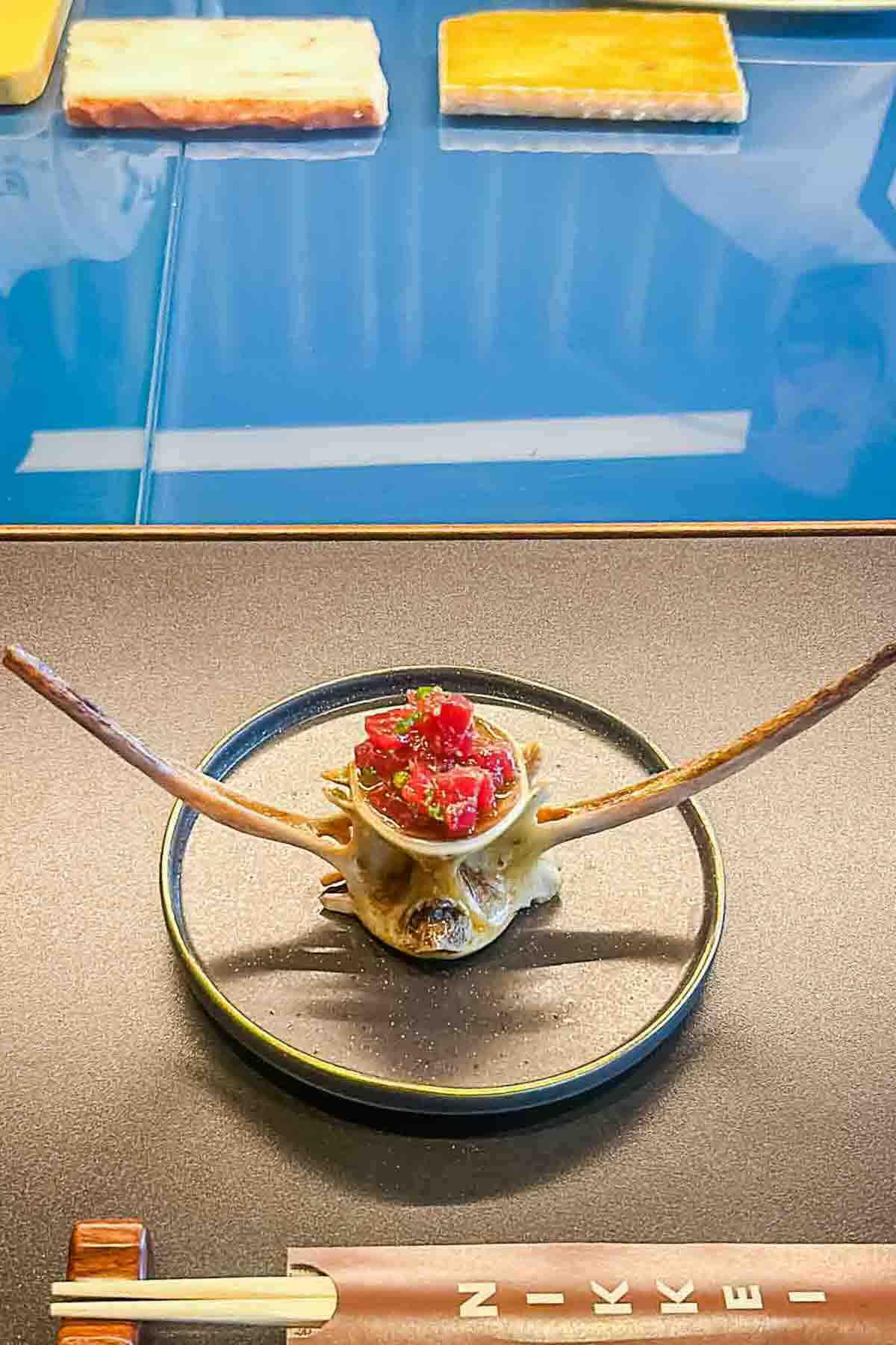 A plate with a seafood dish served in a crab shell with long crab legs, placed beside chopsticks on a dark table.