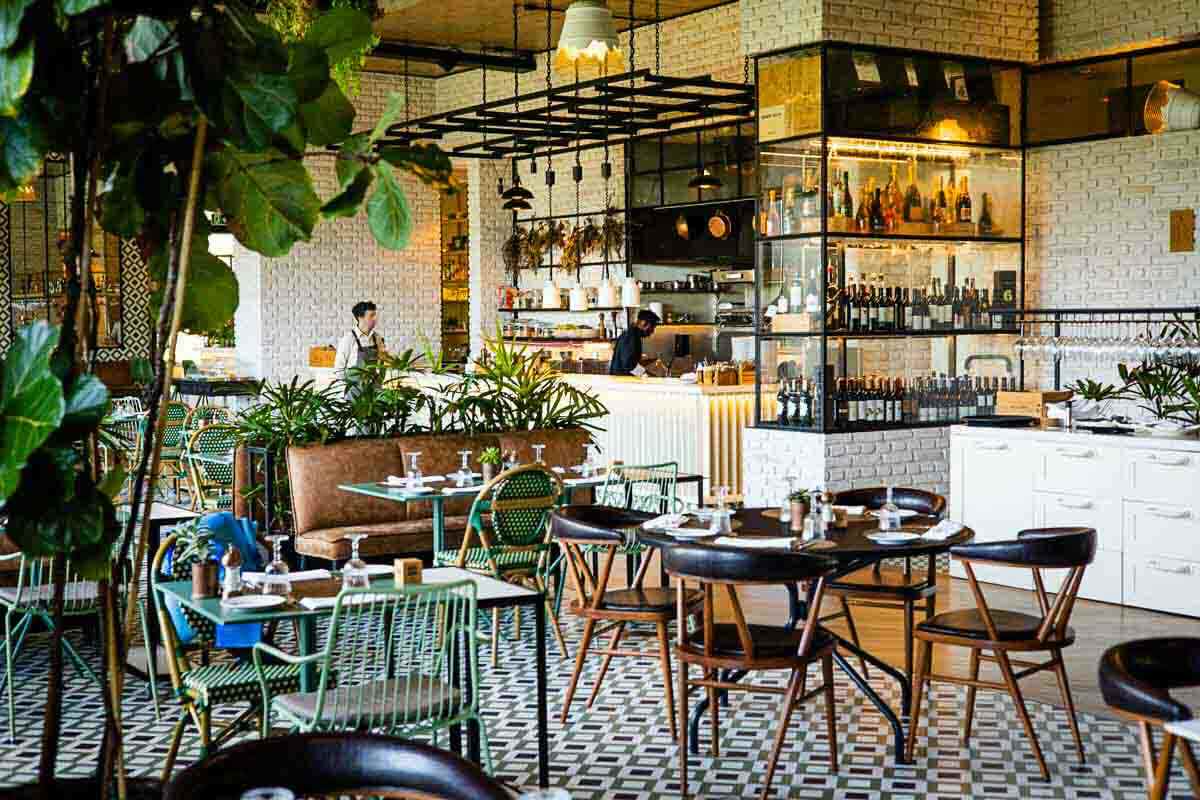 Modern restaurant interior with mixed wooden and green furniture, potted plants, set tables, and an open kitchen with staff working behind the counter.