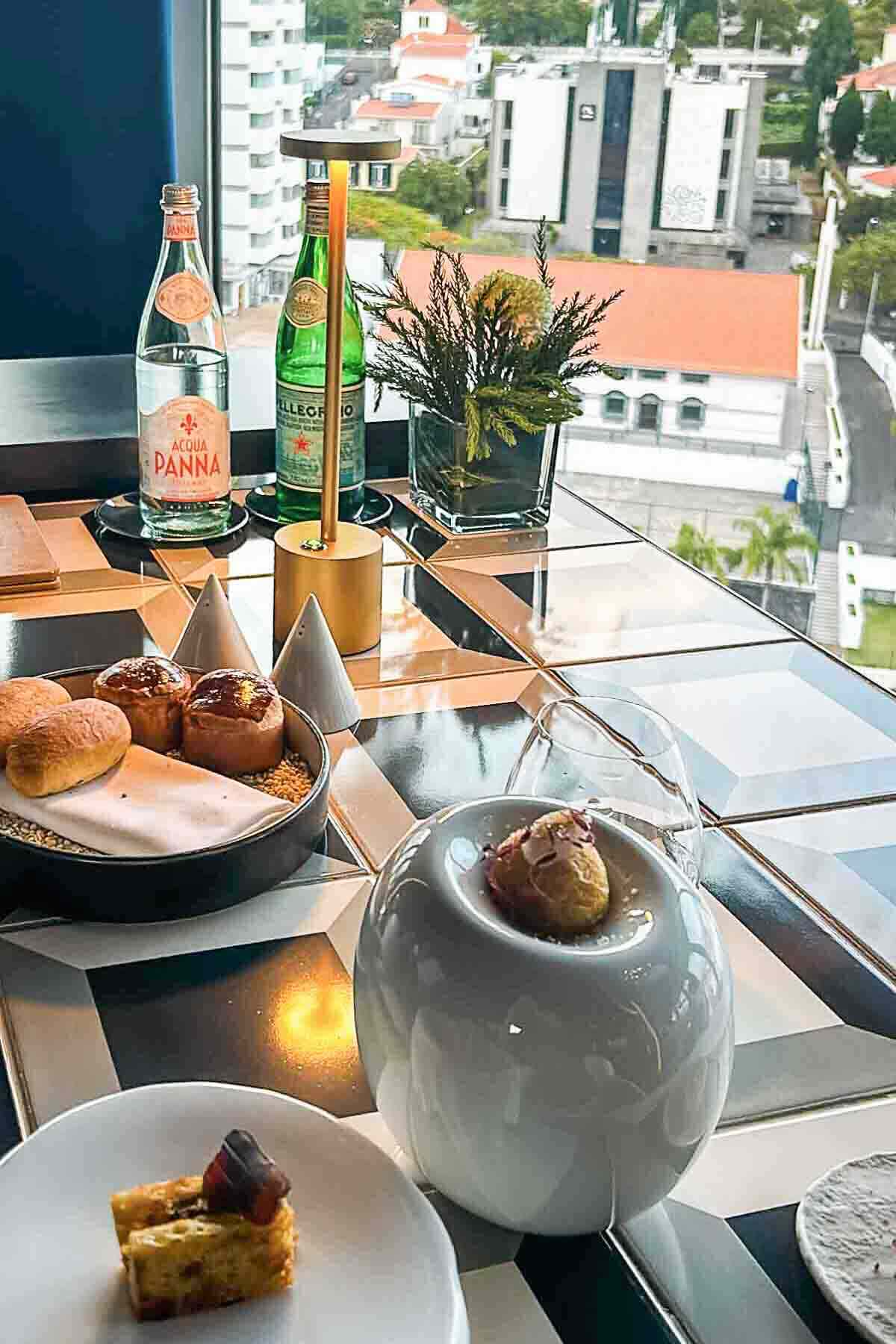 A table set with bottled water, pastries, a bread roll, a plated appetizer, and decorative plants by a window overlooking a cityscape.