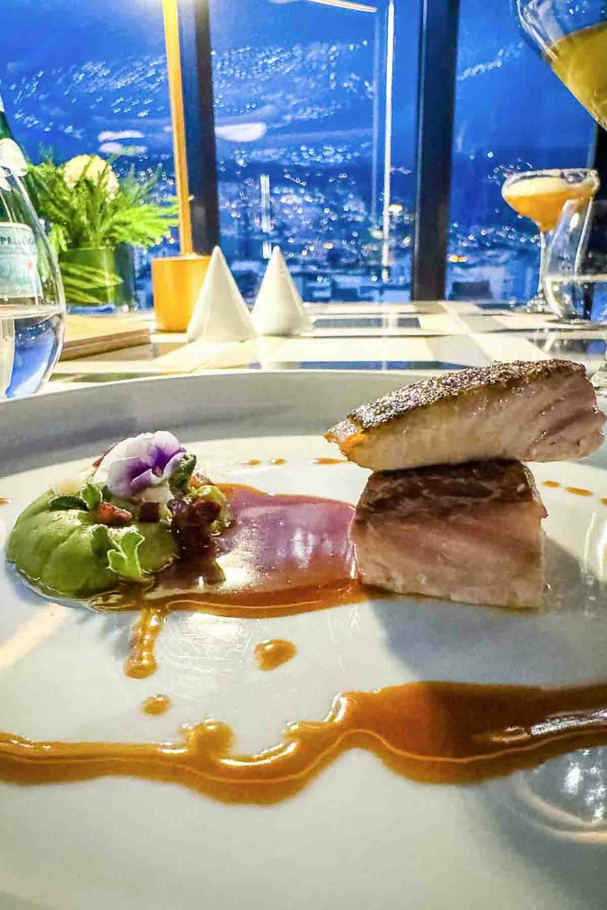 Seared fish fillets stacked on a plate with green puree, edible flower garnish, and sauce, set on a table with city lights visible through a window in the background.